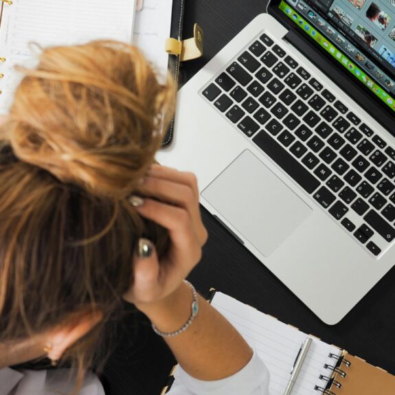 Overhead view of a stressed woman working at a desk with a laptop, phone, and notebooks.